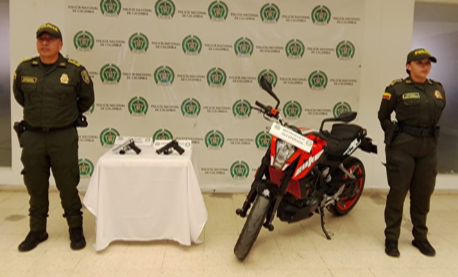 Dos polic&iacute;as una mesa con dos armas de fuego, una motocicleta recuperada