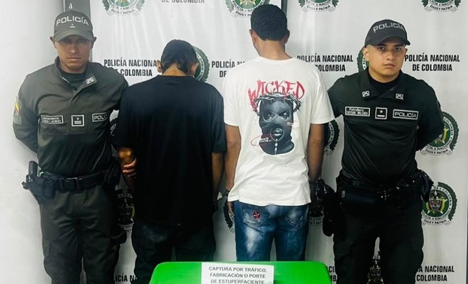 Foto de dos capturados con dos policías a los lados sobre la mesa un papel informado el motivo de su captura