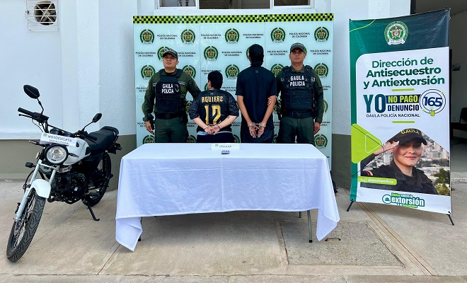 Una motocicleta incautada, un celular incautado sobre una mesa con mantel blanco, dos personas de espaldas capturadas por el delito de extorsión en compañía de dos funcionarios del GAULA de la Policía Nacional brindando seguridad. 