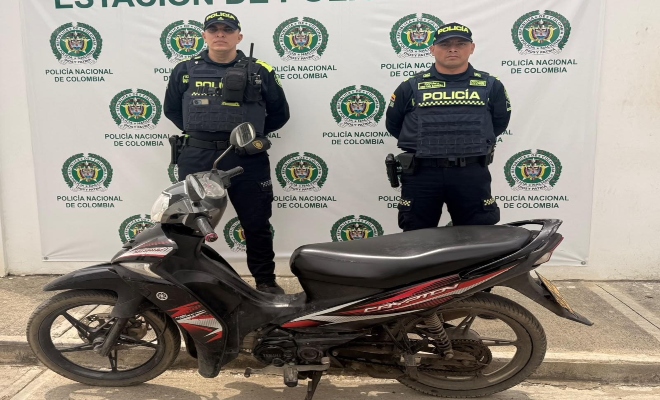Dos funcionarios policiales con una motocicleta recuperada frente a pendón institucional 