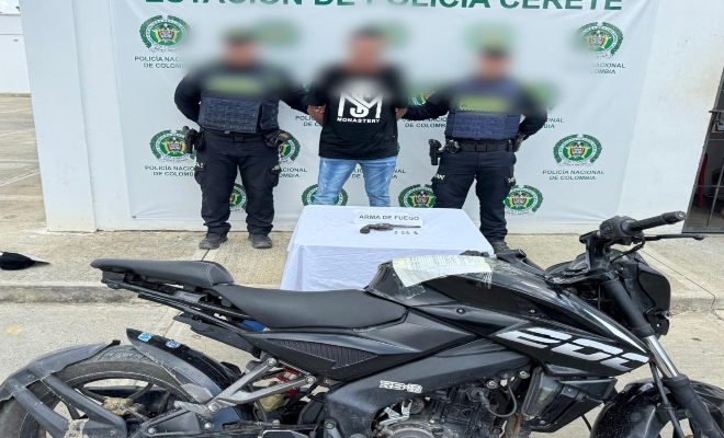 Dos funcionarios junto a un ciudadano capturado, frente a ellos una mesa sobre la cual esta un arma de fuego y delante una motocicleta 