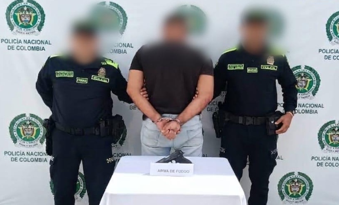 Dos funcionarios policiales con un ciudadano capturado frente a pendón institucional y delante una mesa sobre la cual esta el arma incautada