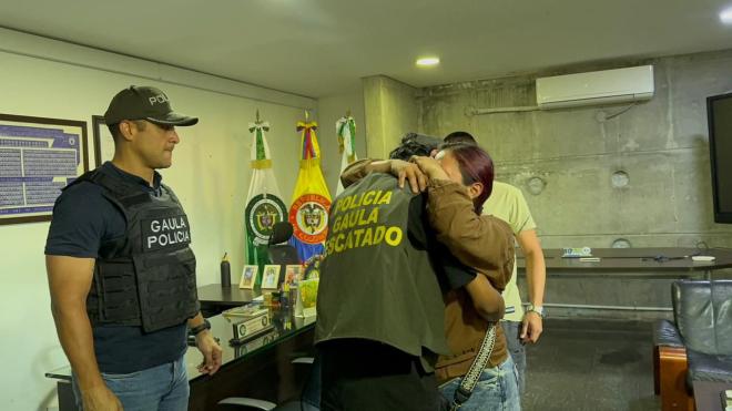 En una oficina institucional, una persona abraza a un familiar, mientras otros funcionarios observan la escena. Al fondo se aprecian banderas y elementos oficiales.