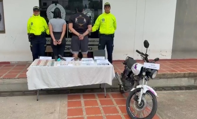 dos Policias y en medio dos personas capturadas, sobre la mesa alucinogenos, una pistola, dinero en efectivo y al lado derecho una motocicleta incautada.