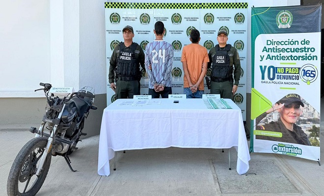 Dos personas de espalda capturadas por el delito de extorsión, elementos incautados sobre una mesa con mantel blanco, dos funcionarios de la Policía brindando seguridad y una motocicleta incautada.