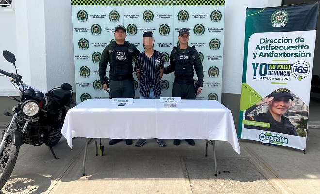 Una persona capturada por el delito de extorsión junto a una mesa con mantel blanco donde están ubicados los elementos incautados, al lado una motocicleta incautada y dos funcionarios del grupo GAULA de la Policía Nacional brindando seguridad