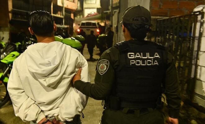 Calle estrecha en zona urbana durante la noche; una persona con chaqueta clara camina con las manos sujetas detr&aacute;s mientras una persona uniformada con chaleco que dice &ldquo;GAULA Polic&iacute;a&rdquo; la gu&iacute;a, con varias motocicletas y un veh&iacute;culo al fondo