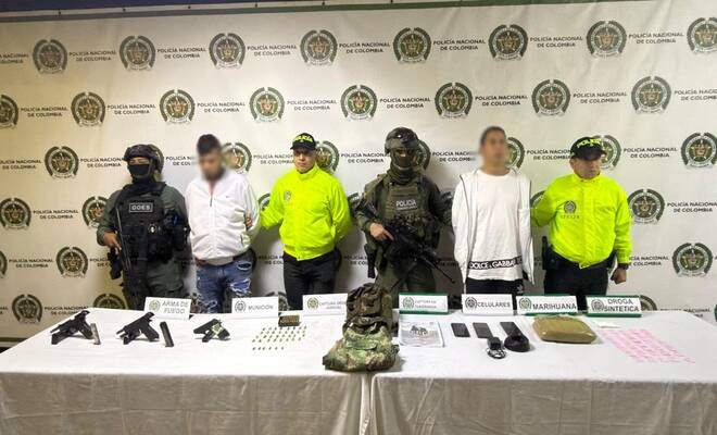 Seis personas de pie frente a un fondo institucional de la Policía Nacional de Colombia; en primer plano hay una mesa con armas, munición, dispositivos móviles y otros elementos identificados mediante rótulos informativos