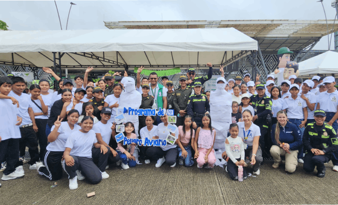 Un grupo de diez policías está de pie acompañados a ambos lados por niños y niñas vestidos con camisas blancas todos se encuentran formados al centro destaca un marco azul y al fondo se observa una carpa blanca