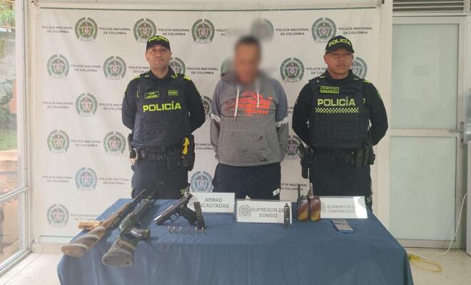 Tres personas de pie frente a un fondo institucional de la Polic&iacute;a Nacional; en primer plano hay una mesa con armas largas, munici&oacute;n y otros objetos exhibidos de forma organizada.