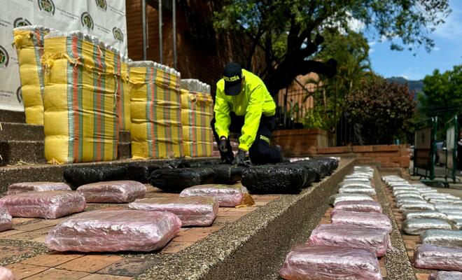 Un investigador de la Policía Nacional en Medellín abre un paquete prensado envuelto en plástico para mostrar que contiene marihuana incautada a un grupo delincuencial. A su alrededor hay varios paquetes similares dispuestos en el suelo.